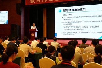 讲台上一位专家正在对着大屏幕讲解智慧体育相关政策，台下坐满了来自全国各地的教育工作者，会议横幅写着教育部智慧体育研讨会字样