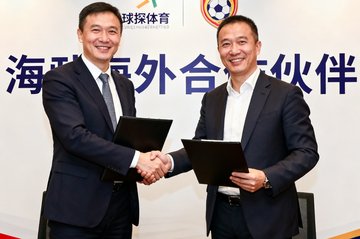 签约仪式现场，双方代表正在交换文件并握手微笑，背景板上印有球探体育和海外合作伙伴的标志，气氛正式而友好